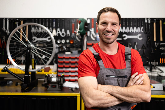 Portrait of experienced serviceman proudly standing in bicycle workshop and holding arms crossed. In background tools and parts.