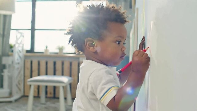little stubborn african american boy draws with markers on the wall in the room