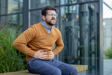 A young man, twisted in pain, sits on a bench near an office building and holds his stomach in his...