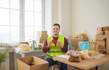 Smiling young volunteer holding red heart in hands sitting at charity center packing donations of foodstuffs in cardboard boxes. Man volunteering in charitable foundation. Humanitarian aid concept