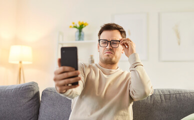 Discontent man focused in smartphone screen, looking through eyeglasses, has bad eyesight, uses...