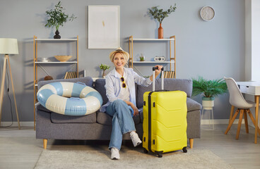 Portrait of a happy woman with suitcase sits on sofa at home living room, ready for summer travel...