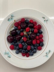 A plate with fresh cherries, blueberries, and raspberries arranged on a white ceramic dish with a...