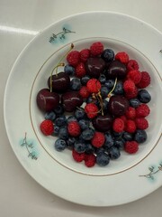 A white plate filled with fresh cherries, blueberries, and raspberries on a plain surface, captured...