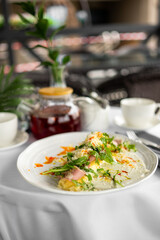 Close-up of a gourmet omelette topped with ham, cheese, and greens on a white plate, served with red tea in a teapot and cups on a table with white tablecloth.