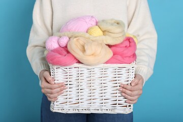 Woman holding wicker basket with colorful felting wool and yarns on light blue background, closeup