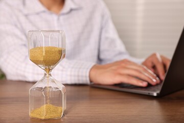 Deadline. Hourglass and man working with laptop at wooden table, selective focus