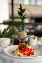 Gourmet breakfast with smoked salmon, poached eggs, and hollandaise sauce served on a white plate with greens. Cozy setting includes teapot, cup, and green plant.