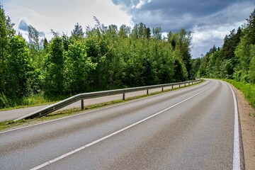 Curved Road in Green Forest Under Cloudy Sky.  The asphalt road is bordered by a metal guardrail...