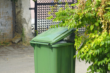 Overflowing green trash bin outdoors on urban street near bushes. Environmental pollution and waste...