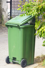 Full green plastic garbage bin on wheels on sidewalk ready for removal by municipal service. Urban waste overflow and outdoor trash disposal. Environmental problem in city with litter and pollution