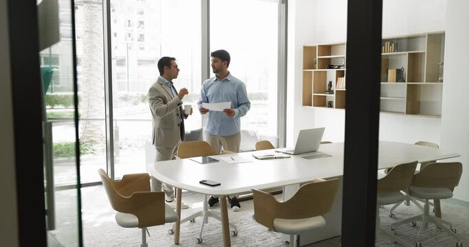 Two businessmen discussing upcoming meeting agenda, standing in office meeting room, one colleague speaking, explaining opinion, solution, idea or point of view to another. Collaborative conversation