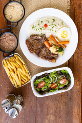 Prato Executivo Brasileiro de bisteca de boi com Arroz, Batata Palha, feijão, farofa e Salada em Mesa de Madeira