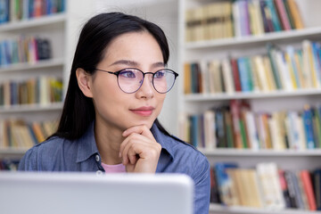 An Asian woman wearing glasses thoughtfully looks into the distance while in a library, with books...