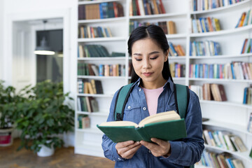 An asian student reads a book in a library setting, surrounded by bookshelves. She wears a backpack...