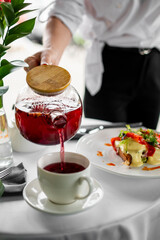 Glass teapot pouring red tea into a white cup on a table with food, creating a cozy, inviting dining scene in natural light.