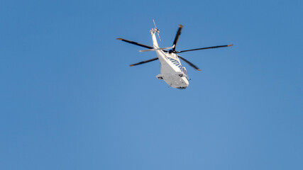Helicopter is flying in the sky. The sky is blue and clear. The helicopter is white and has a blue...
