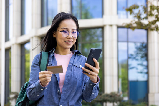 A smiling Asian woman, wearing glasses and holding a credit card, is using her smartphone outdoors near a modern building.