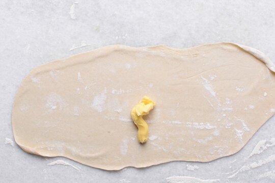 Overhead view of butter being spread on rolled out paratha dough, top view of flaky flatbread dough being covered with ghee, process of making paratha