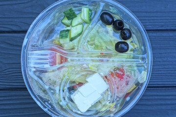 fresh greek salad food in a transparent bowl with sunflower oil and a plastic white fork stands on a black table