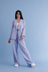 young woman in lilac pantsuit posing confidently on blue background, wearing casual white shoes and...