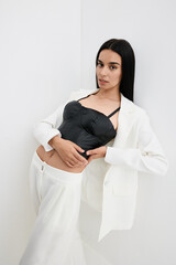 Young woman posing in a stylish black corset and white suit, standing against a plain white...