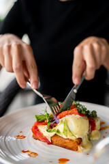 Close-up of a person cutting toast topped with melted cheese, tomatoes, and greens on a white plate with sauce, served as a fresh and appetizing meal.