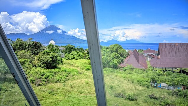 A scenic view of Tidore Island&rsquo;s volcanic mountain from Ternate, North Maluku. Lush green fields, rooftops, and the coast blend under a clear sky&mdash;perfect for nature, travel, and culture themes