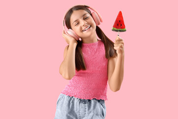 Happy little girl with lollipop in shape of watermelon slice listening music on pink background