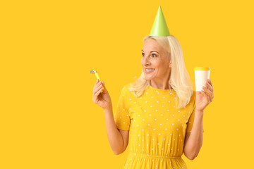 Mature woman with cup of coffee celebrating birthday on yellow background