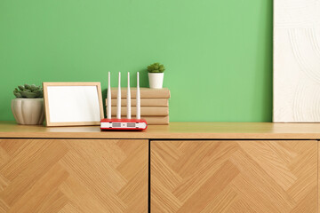 Modern wi-fi router with books, blank frame and plants on commode near green wall