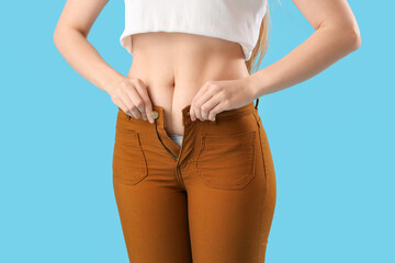 Young woman trying to button tight pants on blue background, closeup. Weight gain concept