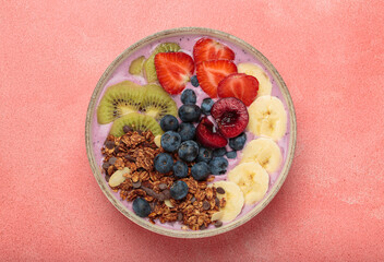 Vibrant and nutritious acai bowl topped with sliced kiwi, strawberries, blueberries, cherries, banana, and granola, a colorful and healthy meal or breakfast on a pink background