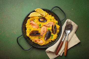 Seafood paella with rice, shrimp, mussels, peas, and lemon wedges served in a black traditional pan on a green rustic background with olives and cutlery