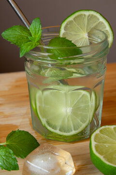 Mojito-like refreshing lemonade with mint, lime and ice in a glass jar with a metal straw on wet wooden cutting board against contrast background, side view