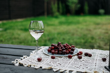 A glass of white wine and a plate of cherries on a table outside on a sunny day. A refreshing glass of white wine alongside a plate of ripe cherries on a table outdoors, ready for enjoyment.