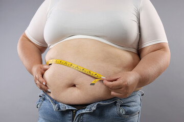 Overweight woman measuring her belly with tape on grey background, closeup