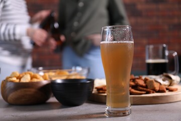 Glass of beer with snacks on table and friends chatting indoors, selective focus