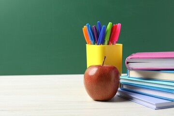 Different school stationery and red apple on white wooden table against blackboard, space for text