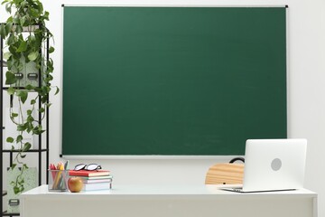 Different school stationery, laptop, glasses and apple on white table near blackboard indoors