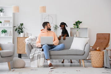 Young man with Dobermann dog using laptop on sofa at home