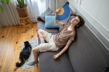 Suffering woman fanning, enduring high temperatures with pet struggling in airless apartment in...