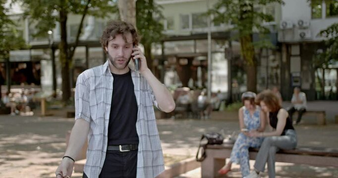 A young man gestures emphatically while talking on his phone in a bustling city square, captured in crisp 4K resolution.