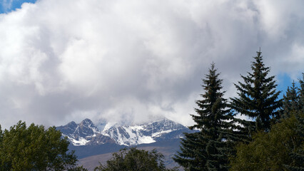 High mountain landscape. Snow-capped peak of rocky mountain in cloud against background of mountain spruce and other trees.