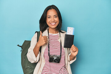 Filipina with camera, tickets, backpack on blue smiling and raising thumb up