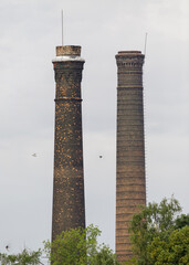 Two aged brick factory chimneys rise above green trees, with birds flying nearby under a cloudy,...