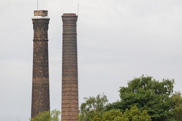 Two tall, weathered brick chimneys from an abandoned industrial site, rising above green trees...