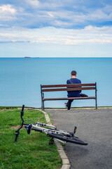 Man with bicycle sitting on bench facing sea. Young man with parked bicycle sitting on bench and...