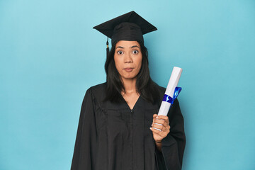 Filipina graduate with diploma on blue studio shrugs shoulders and open eyes confused.
