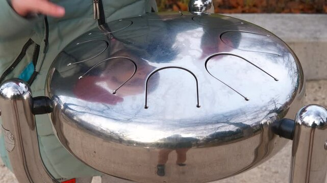 Hands playing melodic notes on outdoor steel tongue drum. Musician's hands creating melodic rhythms on gleaming steel tongue drum, resonating musical tones against natural outdoor background